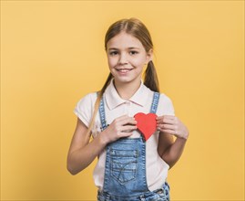 Portrait smiling girl showing red paper cut out heart shape against yellow background
