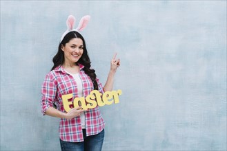 Portrait happy woman with bunny ears head holding easter word pointing finger upward blue background