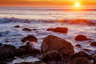 Atlantic ocean sunset with waves and rocks and surfers silhouettes in water at Costa da Caparica