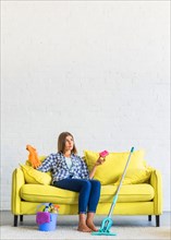 Contemplated young woman sitting yellow sofa holding sponge rubber gloves