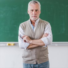 Senior male professor with arms crossed standing against chalkboard