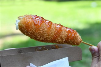 Hand holding Korean style corn dog coated in crispy panko breadcrumbs with hot sauce