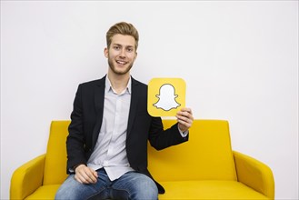 Portrait young man sitting yellow sofa holding snapchat icon