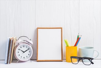 Notebooks alarm clock blank frame pencils holder cup eyeglasses against white wooden wallpaper