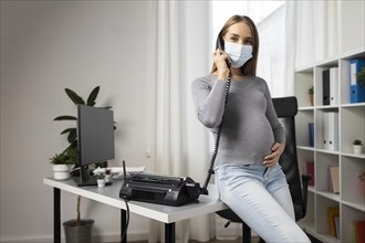 Pregnant businesswoman taking calls office while wearing medical mask