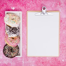 Different type donuts white rectangular box near clipboard with white paper against pink background