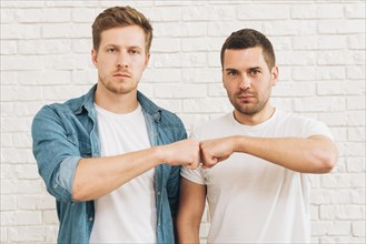 Portrait two male friends bumping fist standing against white brick wall
