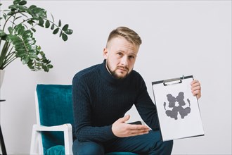 Confident young male psychologist sitting arm chair showing rorschach inkblot test paper