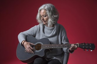 Portrait senior woman sitting chair playing guitar against red background