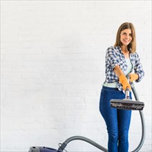 Smiling young woman holding vacuum cleaner front brick wall