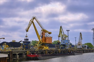 River port cranes with clamshell or griper loading coal to river drag boats or barges moored by pier on cloudy day. Problems of energy production