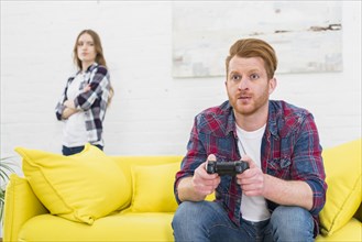 Serious young man playing game with video controller with her girlfriend standing background