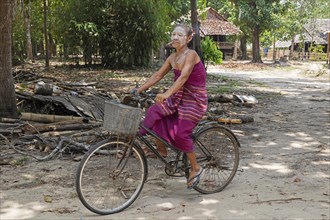 Old Burmese woman wearing thanaka and smoking a cigar while cycling in Kayin village near Hpa-an