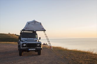 Off-road four-wheel drive vehicle with rooftop tent camping along the Baltic Sea at sunrise in summer