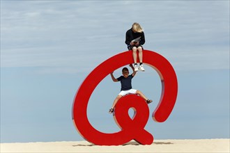 Children sitting on an oversized red at-sign @ the beach