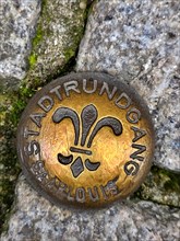 Brass-coloured metal medal embedded in a cobblestone street. The medal is circular and has a fleur-de-lis symbol in the centre. The medal is inscribed with the words Stadtrundgang and Saarlouis around...