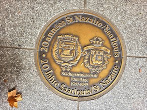 Bronze plaque embedded in a pavement. The plaque is in German and reads 20 years of St Nazaire Saarlouis town twinning jumelage 1969-1989. The plaque has two coats of arms. Saarlouis