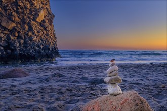 Small balance stone stack and big natural rock lit by sunset sun on sandy beach. Tidal waves in background