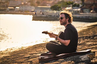 Hipster street musician in black playing electric guitar in the street sitting on pier embankment on sunset