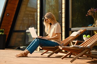 Woman using laptop sitting in a folding wooden chair on the terrace of wooden cabin