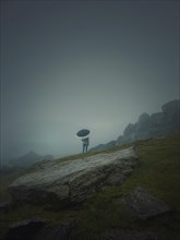 Rear view of a lonely man with umbrella stands on a rocky hill covered by haze. Moody and emotional scene with a lone stranger silhouette under the rain