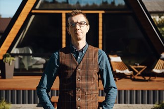 Outdoor portrait of middle aged man in jean shirt and wool vest with tiny house on background