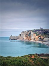 Idyllic view to Etretat coastline with the famous Notre-Dame de la Garde chapel on the Amont cliff. Shore washed by Atlantic ocean waters