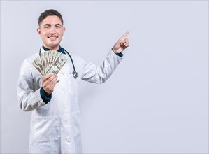 Happy latin doctor holding money and pointing to side. Handsome smiling doctor showing money and pointing side