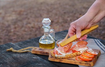 Woman putting slices of iberian ham on a slice of bread with tomato and olive oil on a wooden table in the countryside