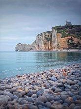Sightseeing view to Etretat coastline with the famous Notre-Dame de la Garde chapel on the Amont cliff. Pebble beach washed by Atlantic ocean waters