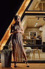 Happy woman in transparent beach dress with a glass of wine on terrace of log cabin in the evening