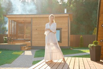 Dreamy barefoot woman in long white dress standing on terrace with hot tub and sauna on background