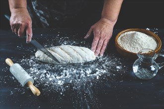 Woman cutting with a knife a flour dough to make homemade bread on a black wooden table with flour