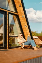 Mid adult woman scrolling social media on laptop while laying in a folding wooden sun lounger on the terrace of a-frame cabin