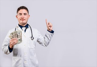 Young latin doctor showing money and pointing side. Cheerful doctor holding money and pointing a advertising