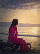 Carefree young woman aesthetic portrait sitting relaxed on the sunbed at the beach looking at the sunrise above the sea. Beautiful seaside dawn scene. Summer holiday