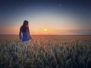 Young woman in the golden wheat field at sunset. Beautiful twilight scenery under the summer sun and crescent moon with starry sky