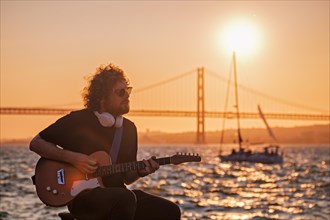 Hipster street musician in black playing electric guitar in the street on sunset with bridge and yacht boat in background