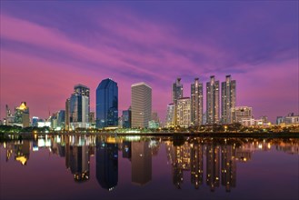 Beautiful landscape with modern high-rise buildings reflecting in calm lake waters at colorful sunset. Benjakitti Park