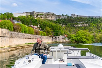 Man steering a houseboat on the river Doubs in front of the World Heritage Site of the Citadel of Besancon