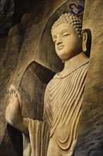 Statue of standing Buddha in monk clothes carved in limestone cave and lit by light. Right hand in adhay mudra gesture. Religion