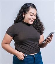 Smiling latin girl using cell phone isolated. Cheerful young woman with curly hair texting on cell phone isolated