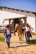 Two cowgirl women coming out smiling pulling a horse out of a stable