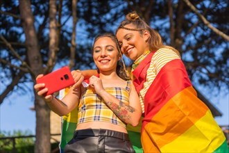 A closeup shot of two young Caucasian hugging females with LGBT pride flag taking selfie outdoors