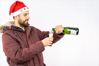 Young very happy Caucasian man with red Christmas hat pouring himself a glass of champagne on a white background