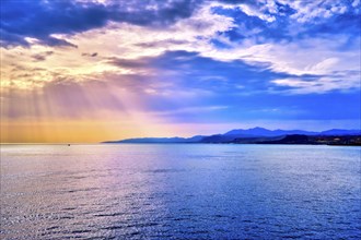 Colorful seascape with colorful clouds and bright sun rays cast over calm sea waters and hazy distant shores. Sunrise or sunset in Mediterranean sea
