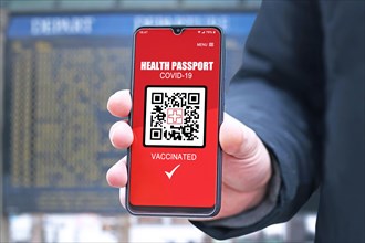 Corona virus vaccine passport on mobile phone device held by hand in front of air port or train station departure board to allow vaccinated people to travel