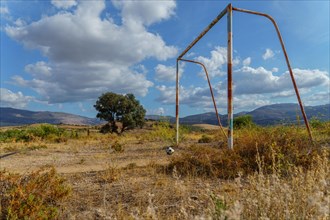 Old rusty soccer goal in an overgrown field with a broken soccer ball in front of it