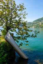 Hanging Boat and a Tree on the Waterfront to Lake Lugano with Mountain in a Sunny Summer Day in Ticino