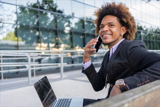 African american young businessman talking to the mobile while working on remote sitting on a bench outdoors